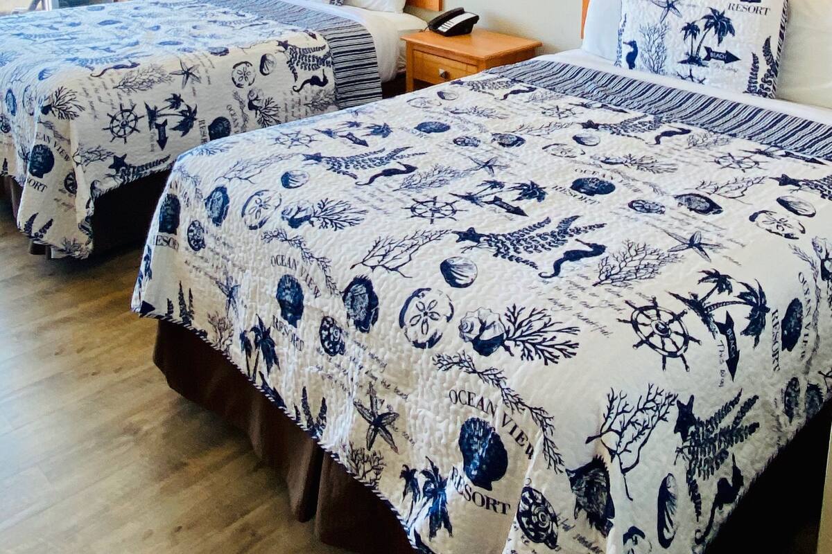 Habitación de hotel con dos camas, cada una con colcha de motivos náuticos. Cuenta con cabecera de madera, mesita de noche con teléfono y ventana con cortinas.