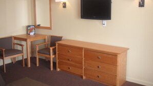 A hotel room with a TV mounted on the wall, a wooden dresser, two chairs, a table, and two wall-mounted lamps. - Oceanview Inn (Emerald Isle)