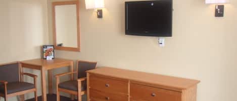 Une chambre d’hôtel équipée d’un téléviseur fixé au mur, d’une commode en bois, de deux chaises, d’une table et de deux lampes fixées au mur.