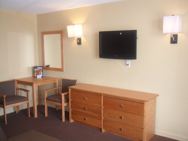 A hotel room with a TV mounted on the wall, a wooden dresser, two chairs, a table, and two wall-mounted lamps.