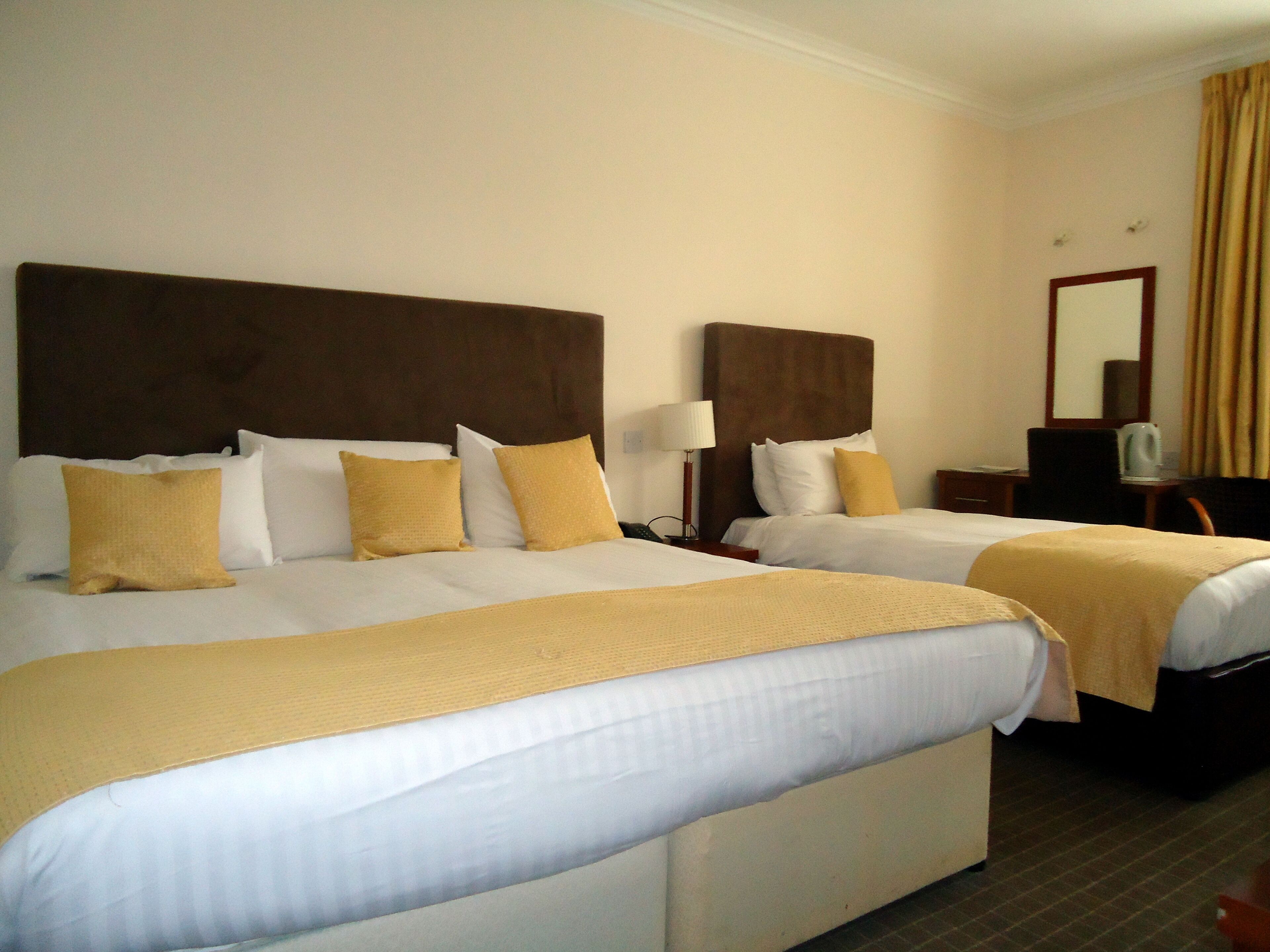 A hotel room with two beds, each with a headboard, white bedding, and a yellow throw blanket. There is a nightstand with a lamp and a small table with a cup and a kettle.