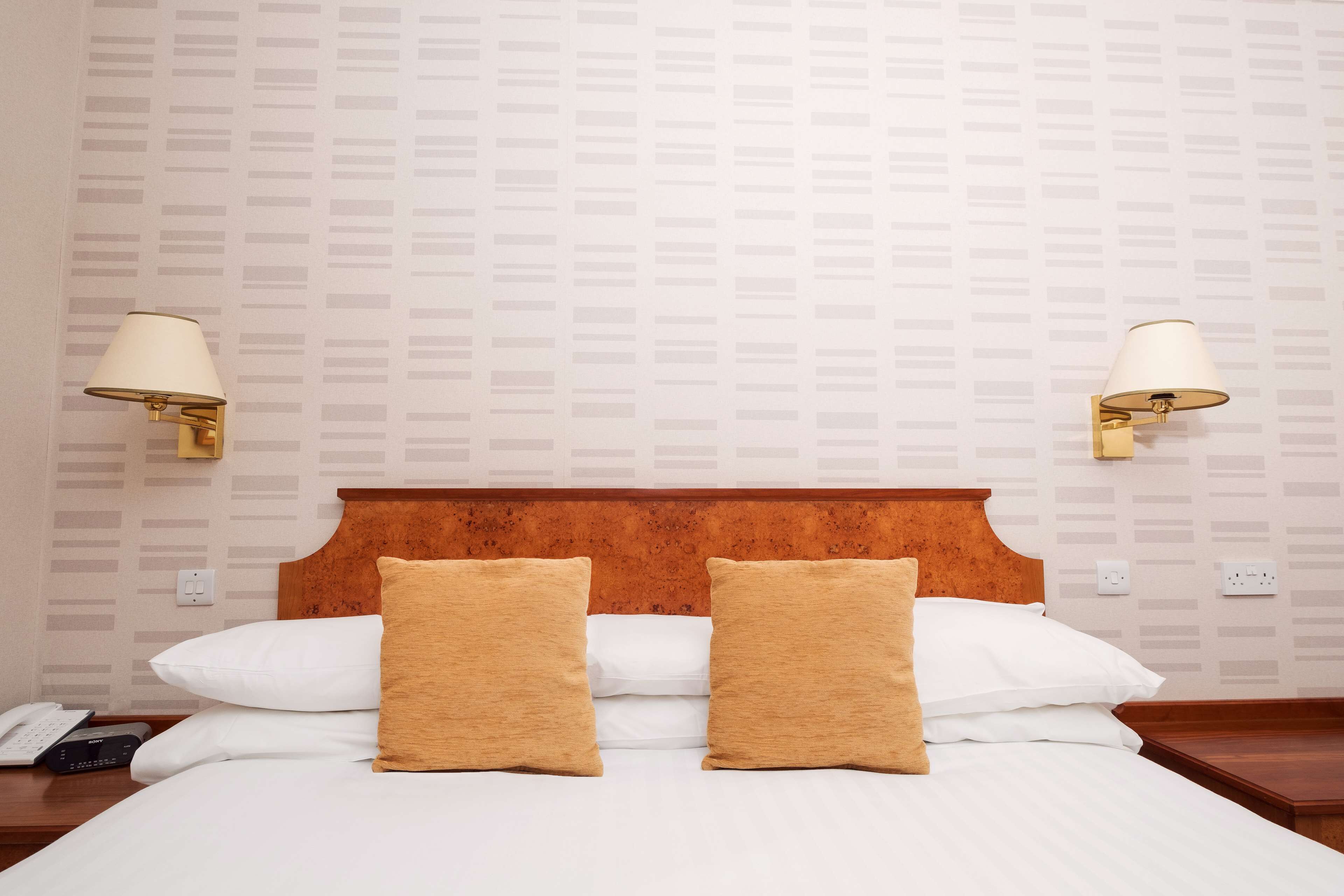 Hotelkamer met een houten hoofdeinde, twee witte kussens, twee beige kussens en twee wandlampen.