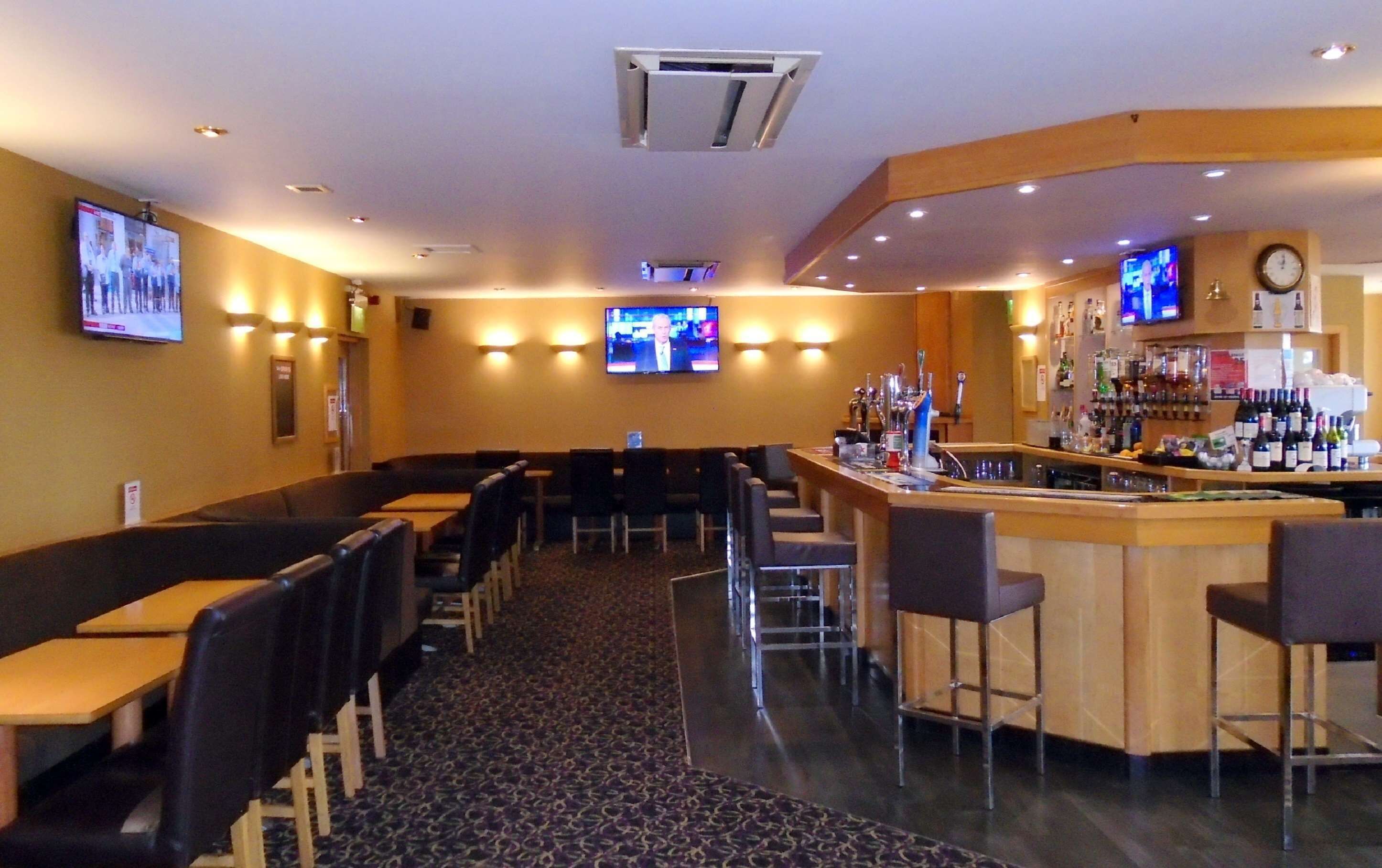 A restaurant with multiple tables and chairs, a bar area with a counter and stools, and two televisions mounted on the walls.