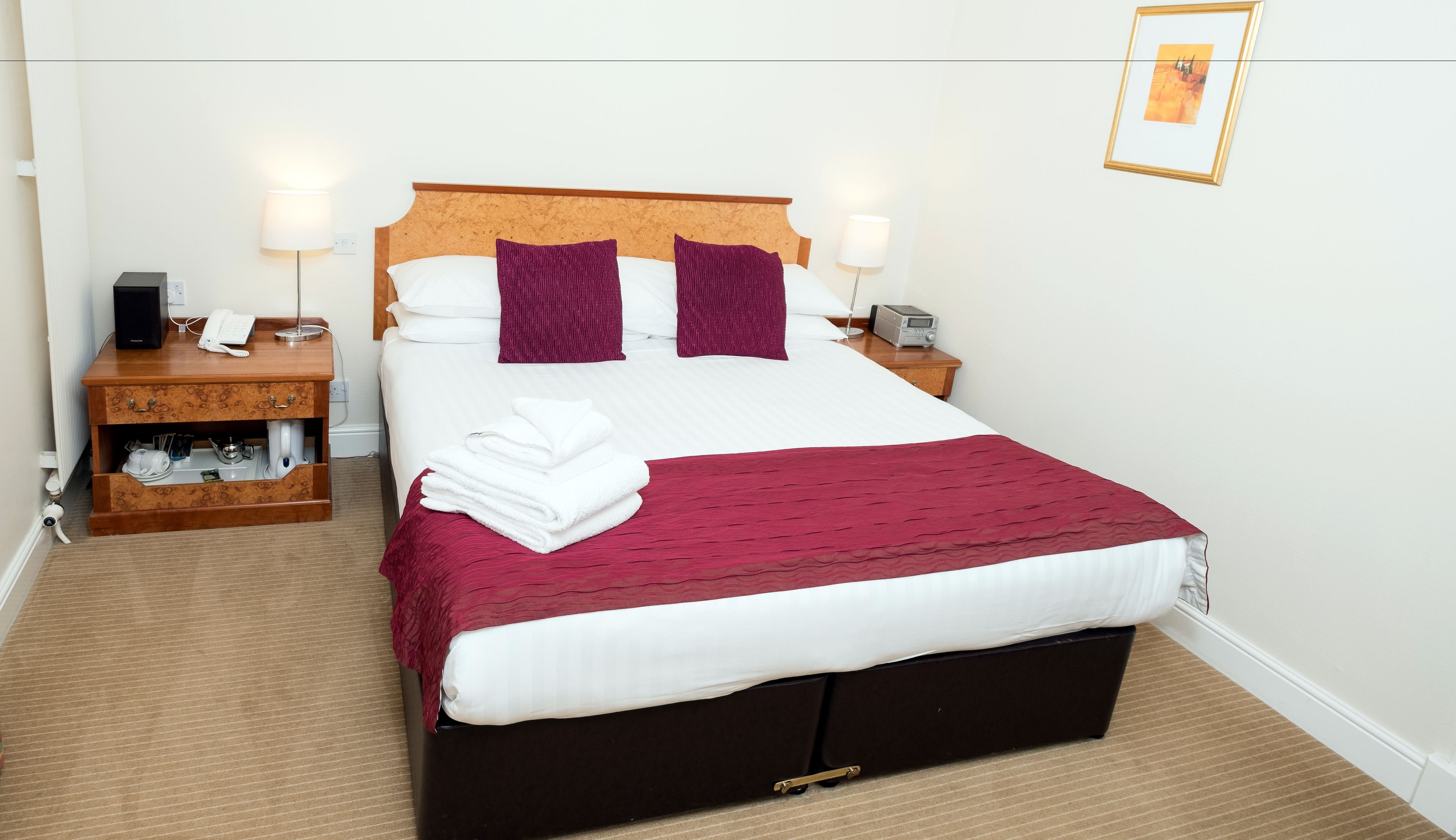 Una cama bien hecha con tablero de madera, ropa blanca y dos almohadas moradas. Hay una manta roja sobre la cama, una mesita de noche de madera con una lámpara y un radio al lado de la cama.