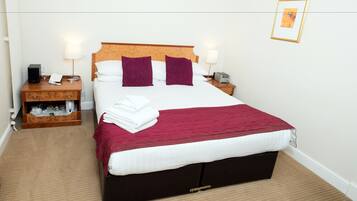 Una cama bien hecha con tablero de madera, ropa blanca y dos almohadas moradas. Hay una manta roja sobre la cama, una mesita de noche de madera con una lámpara y un radio al lado de la cama.