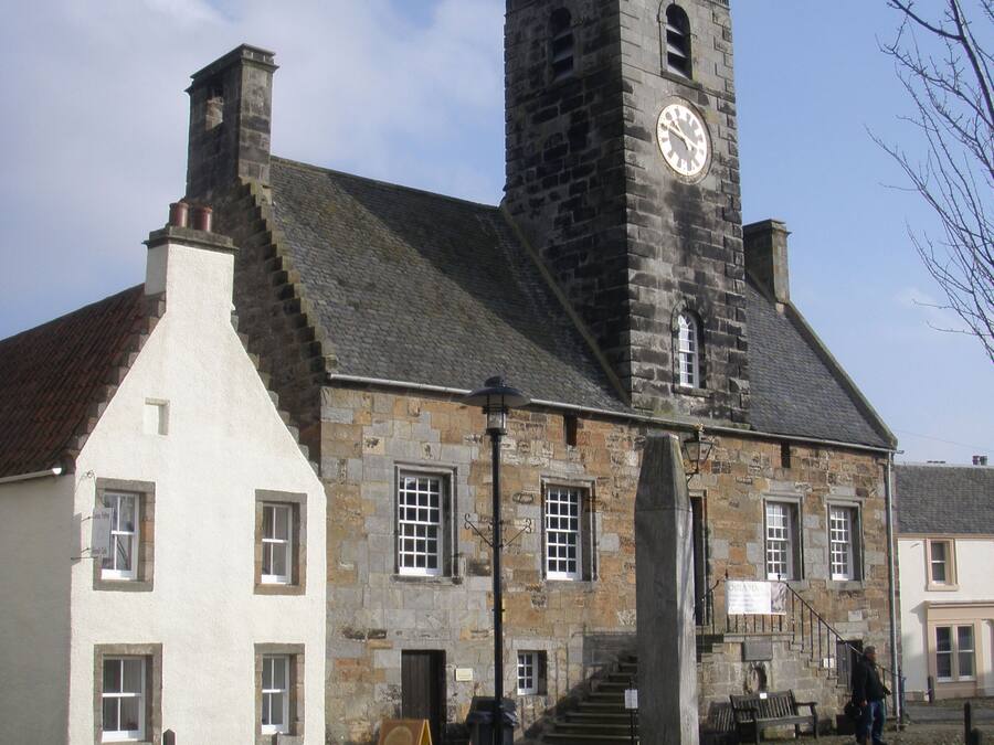 Un edificio histórico de piedra con una torre del reloj, un patio empedrado y una pequeña fuente.