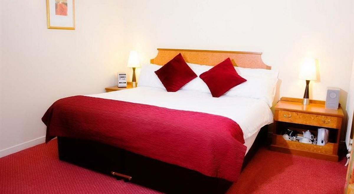 A neatly made bed with a red bedspread and pillows, a wooden nightstand with a lamp, and a small shelf with items.