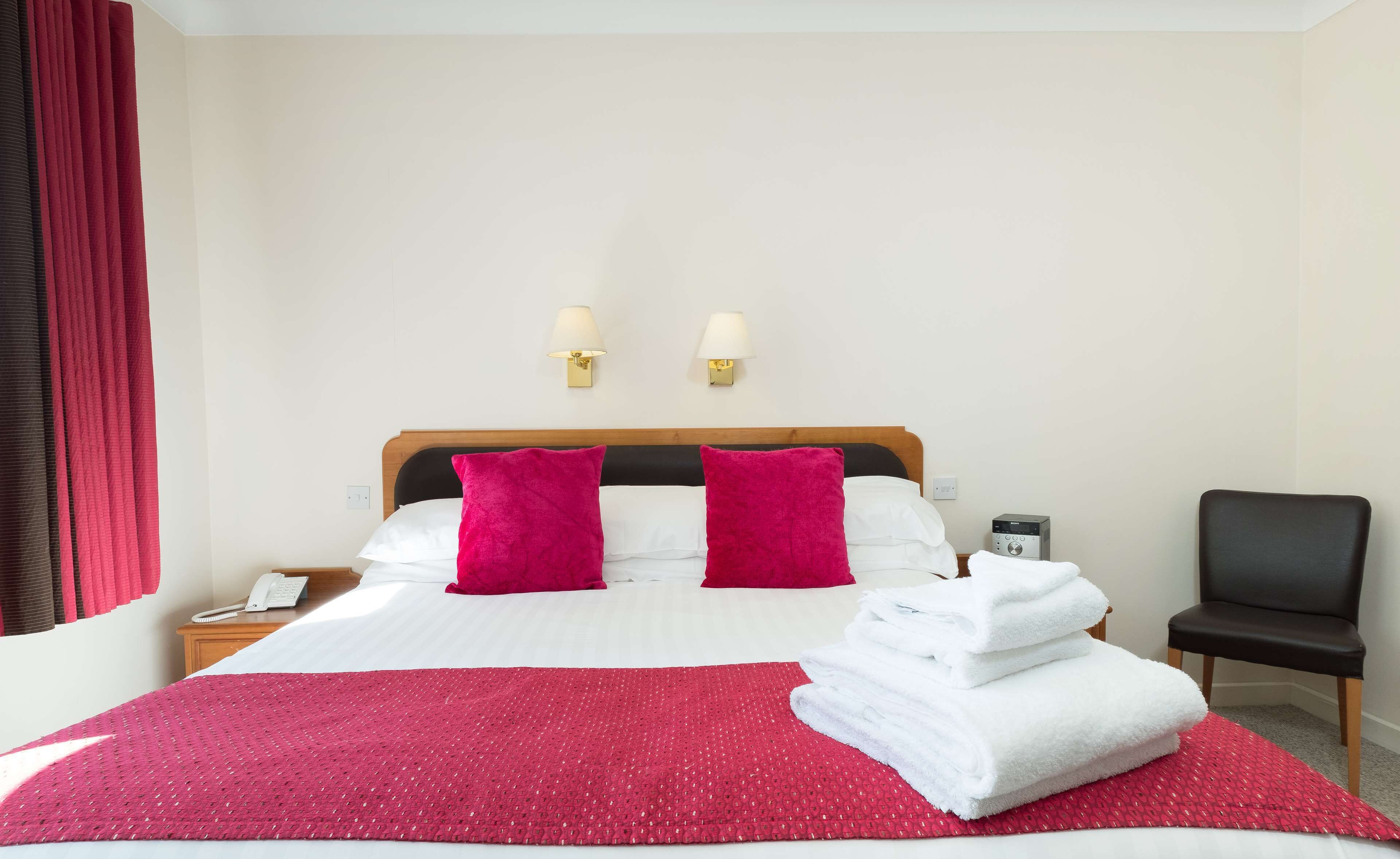 Habitación de hotel con cama, dos lámparas de pared, una silla y un teléfono sobre una mesita de noche.