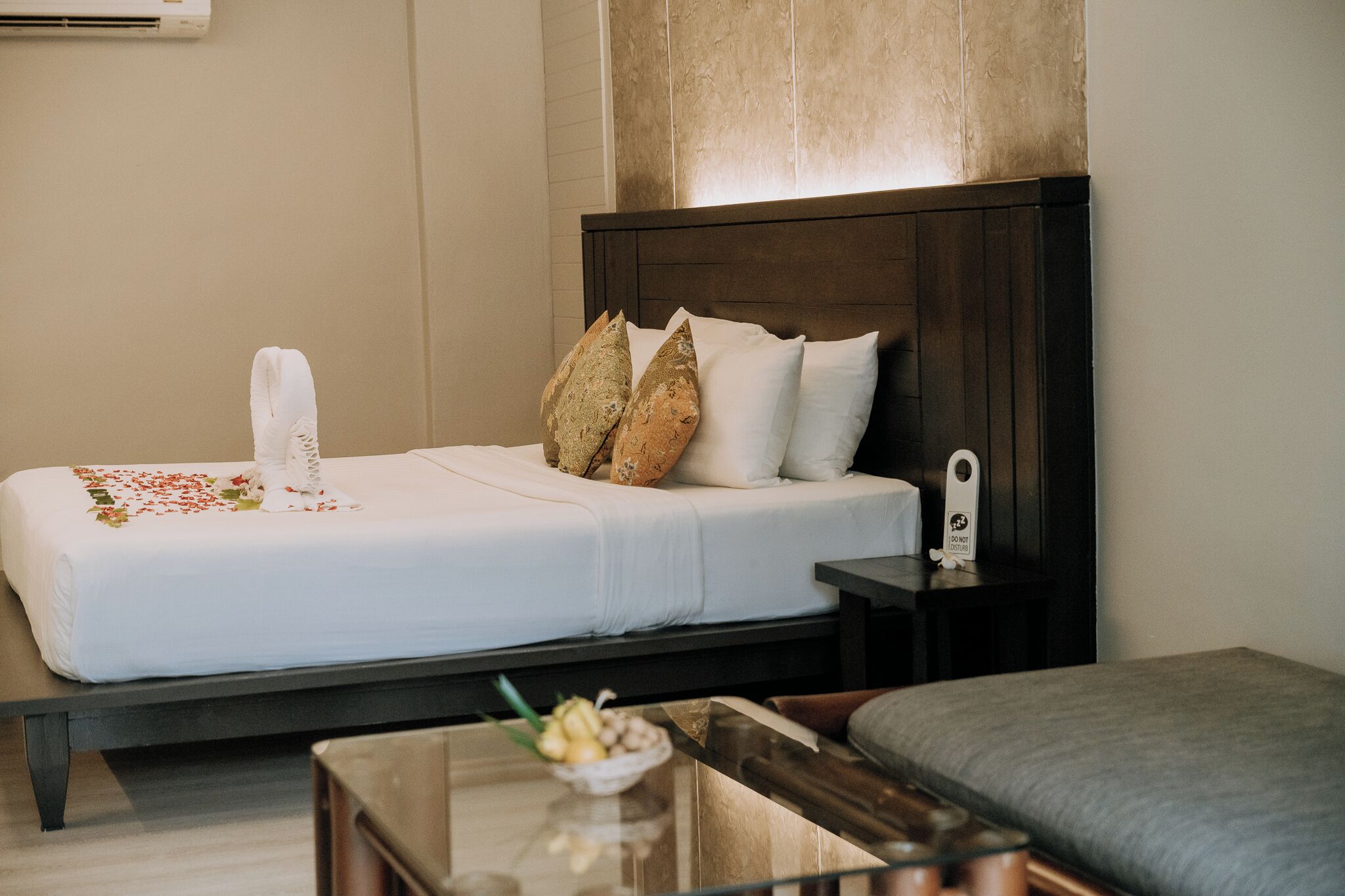 Una cama bien hecha con almohadas, una mesita de noche con un reloj y una pequeña mesa con un cuenco de fruta.