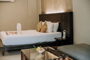Een net opgemaakt bed met kussens, een nachtkastje met een klok en een klein tafeltje met een schaal fruit.