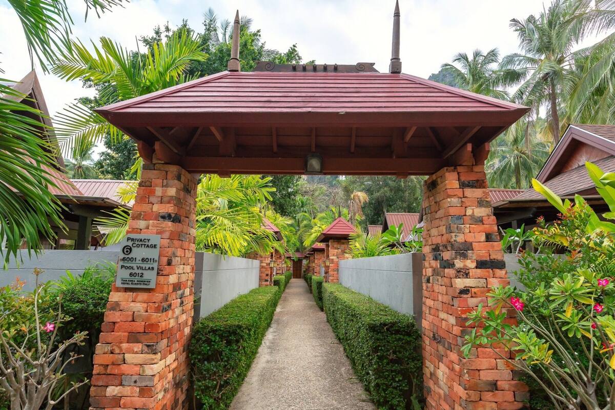 A pathway leading to a brick structure with a red roof, surrounded by lush greenery and palm trees.