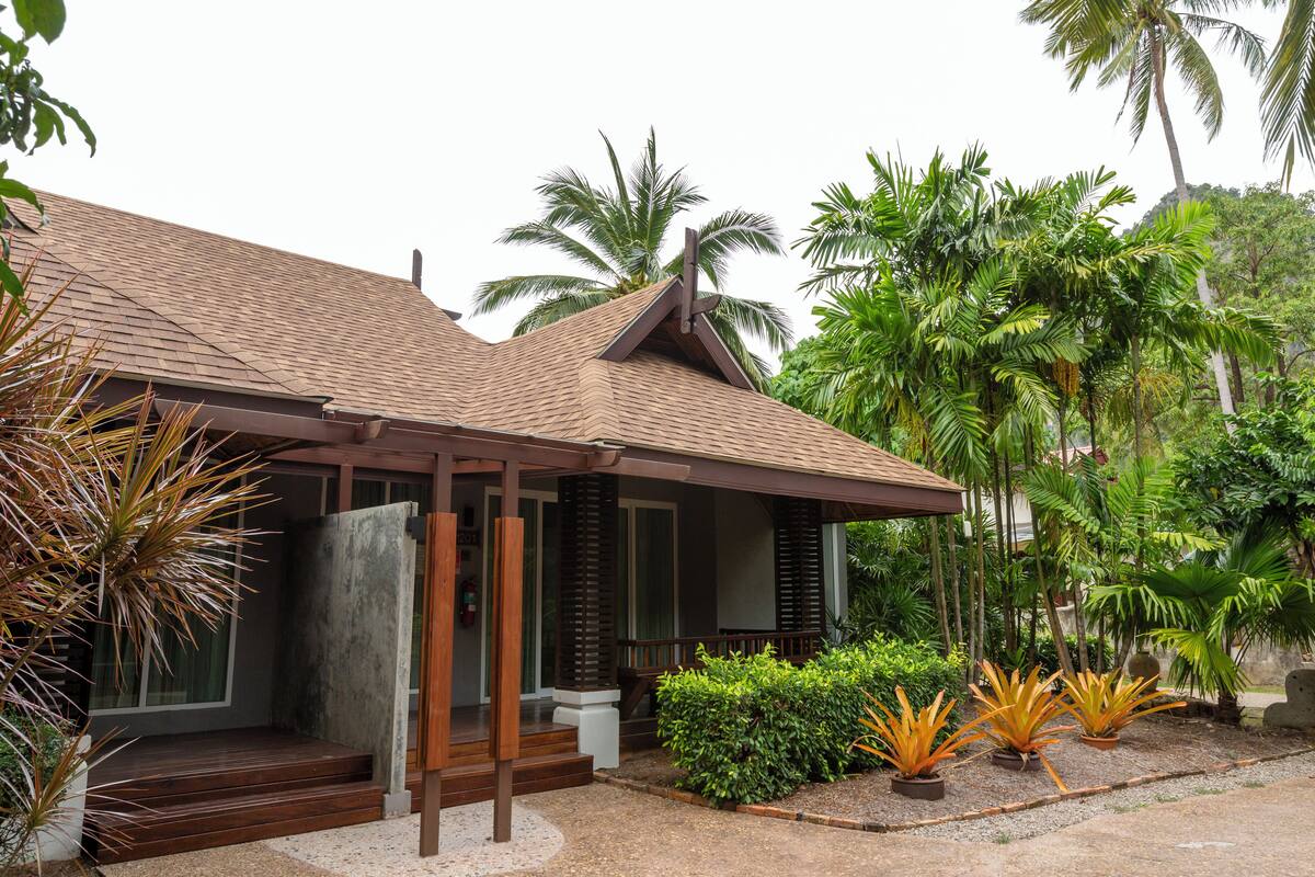 A modern house with a tiled roof, wooden porch, and a garden with palm trees and plants.