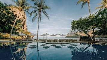 Una piscina con sillas de descanso y sombrillas, palmeras y vista al mar.