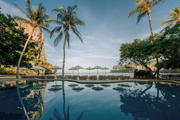 A pool with lounge chairs and umbrellas, palm trees, and a view of the sea.