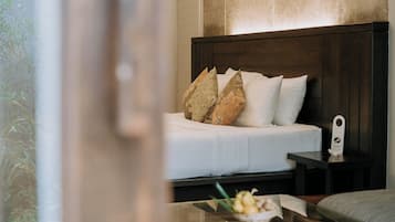 Una cama bien hecha con ropa blanca y almohadas decorativas, una mesita de noche con una lámpara y una pequeña mesa con un cuenco de fruta.