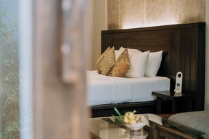 Una cama bien hecha con ropa blanca y almohadas decorativas, una mesita de noche con una lámpara y una pequeña mesa con un cuenco de fruta.