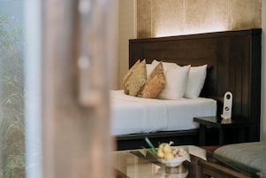 Een netjes opgemaakt bed met witte beddengoed en decoratieve kussens, een nachtkastje met een lamp, en een bijzettafeltje met een schaal fruit.