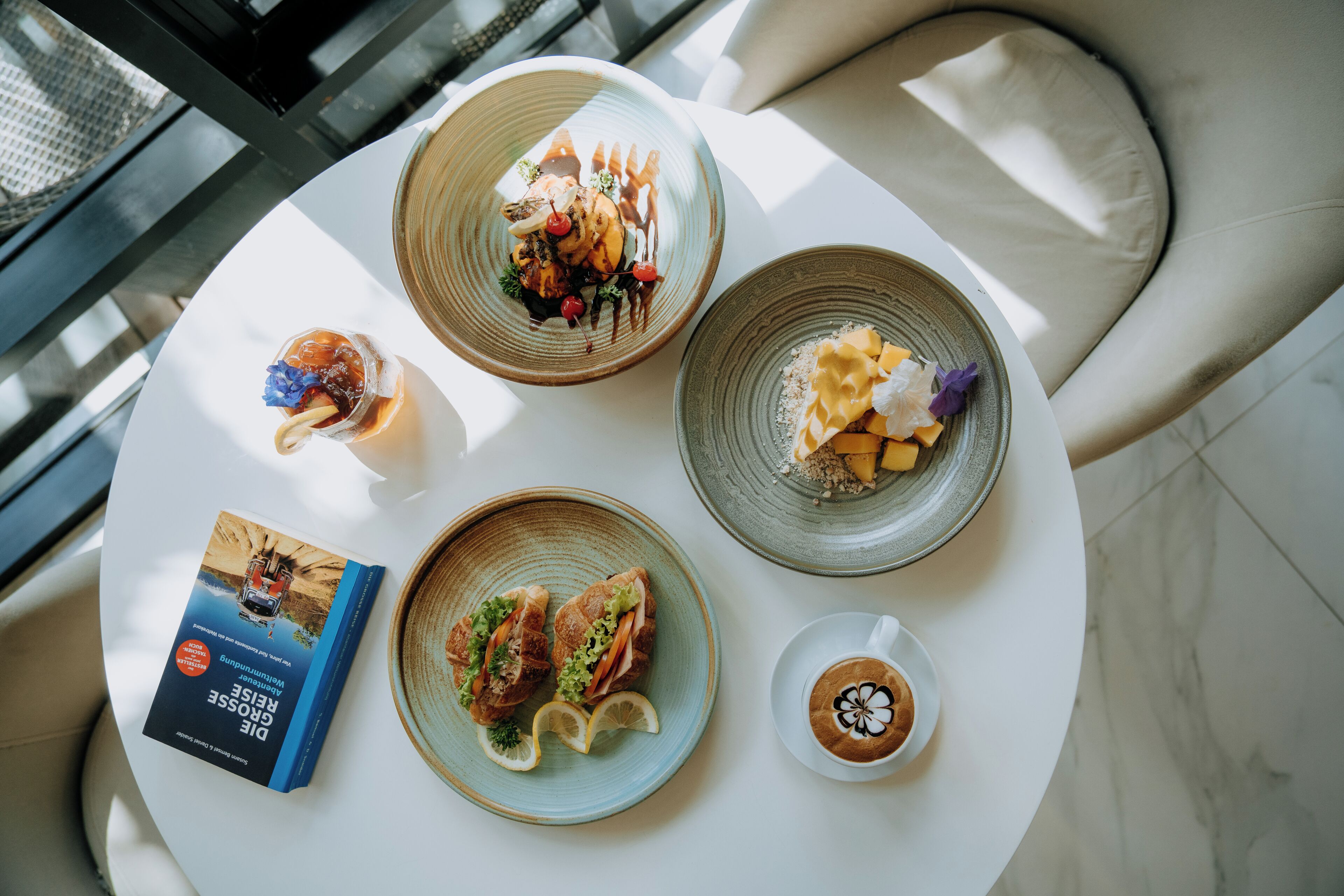 A table with a book, two plates of food, a glass of iced beverage, and a cup of coffee.