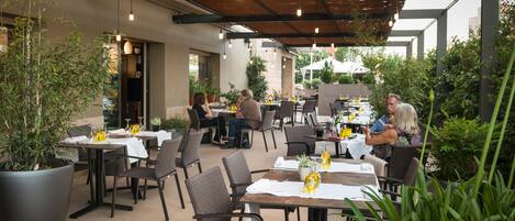 An outdoor dining area with multiple tables and chairs, surrounded by potted plants and a wooden ceiling.