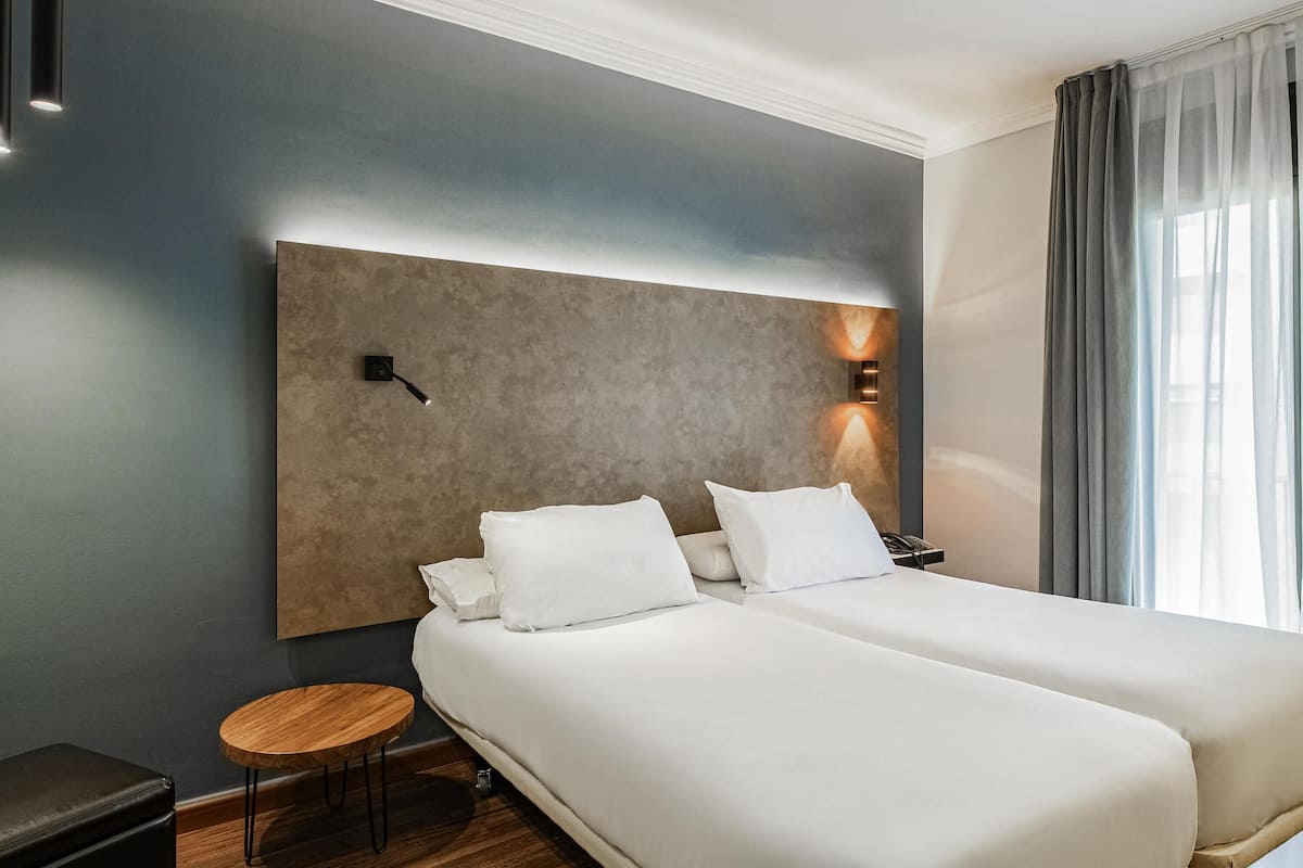 A hotel room with two beds, a small round table, a nightstand, and a modern headboard with wall-mounted lights.