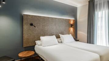 A hotel room with two beds, a small round table, a nightstand, and a modern headboard with wall-mounted lights.
