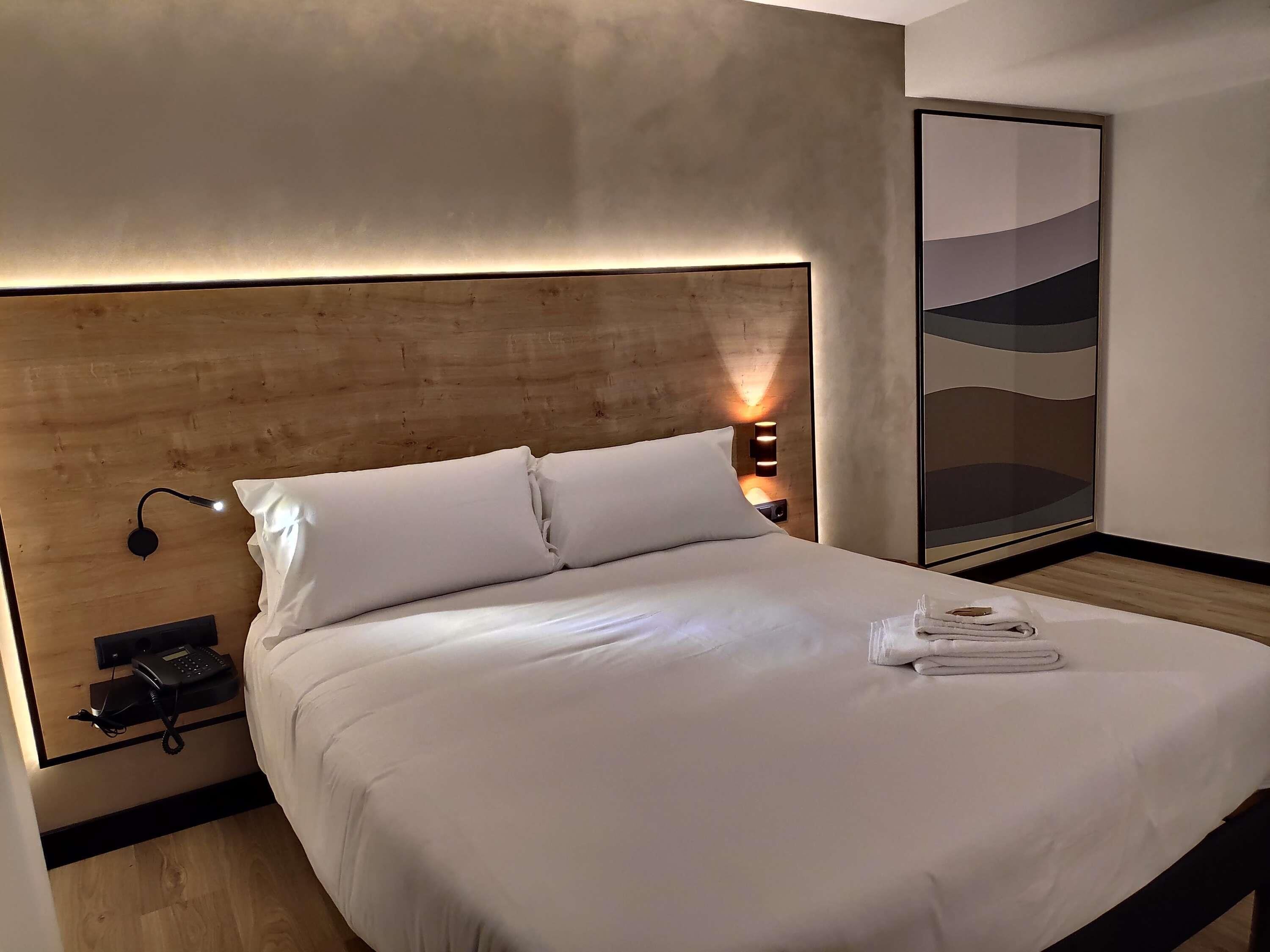 A neatly made bed with white linens, a bedside phone, and a wall-mounted lamp.