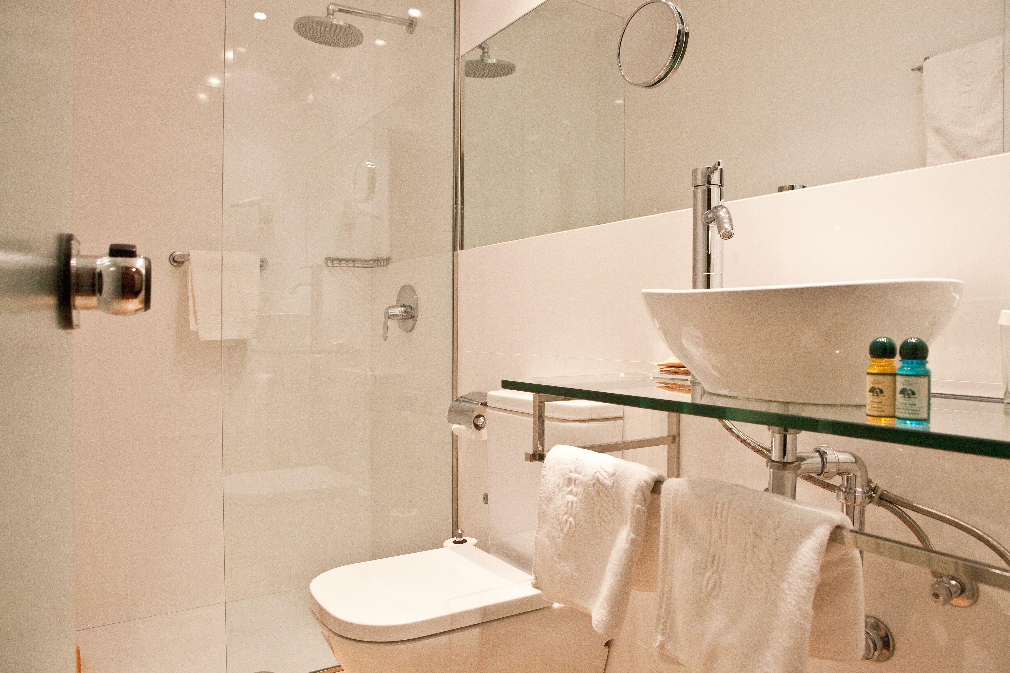Un bany modern amb plat de dutxa de vidre, vàter blanc i un lavabo de vidre amb mirall a sobre.
