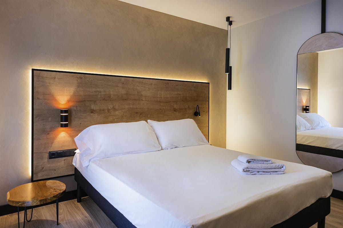 A modern hotel room with a large bed, a wooden headboard, a small side table, and a mirror on the wall.