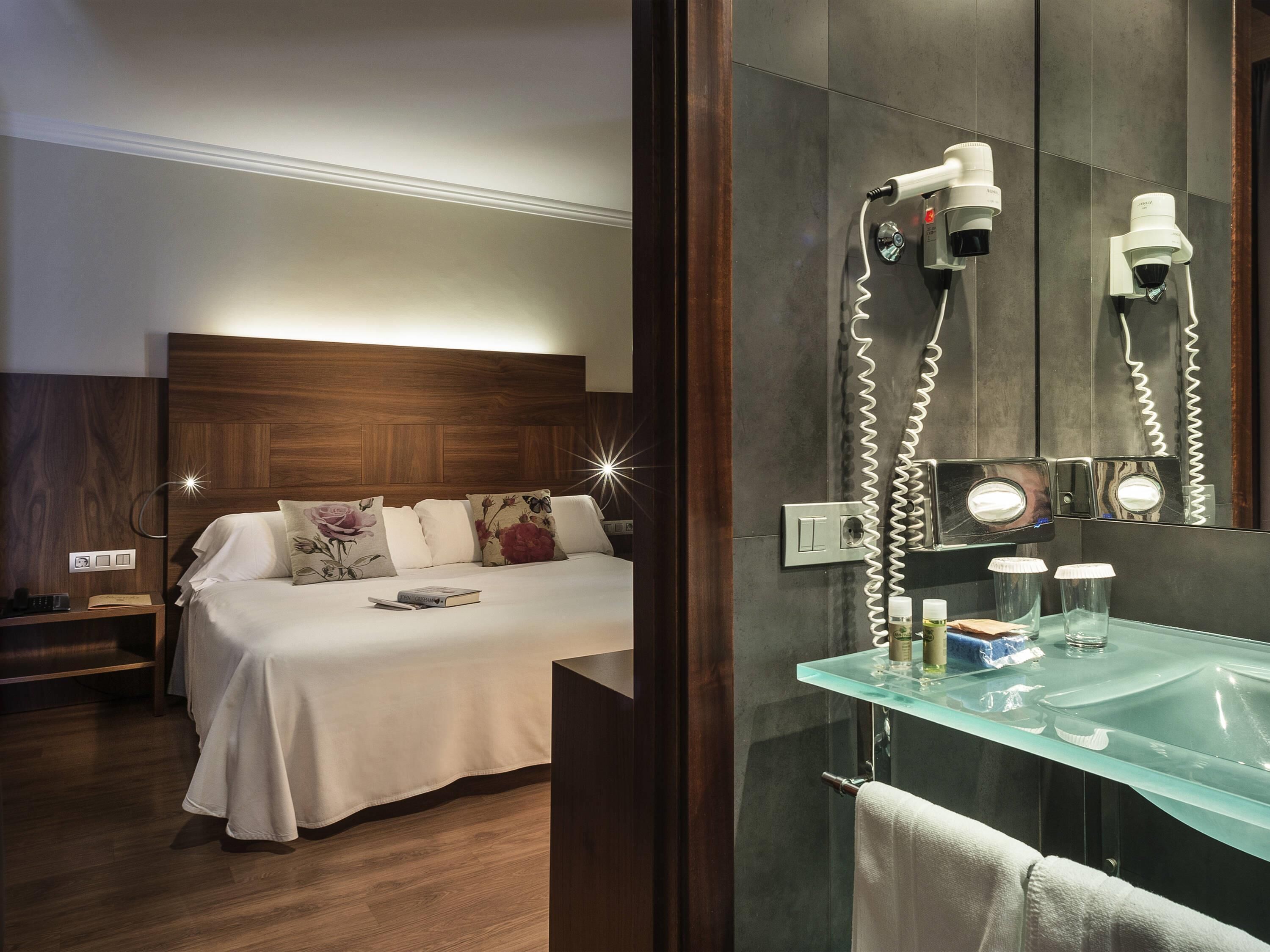 A hotel room with a bed, bedside table, nightstand, and a bathroom with a glass sink, mirror, and towel rack.