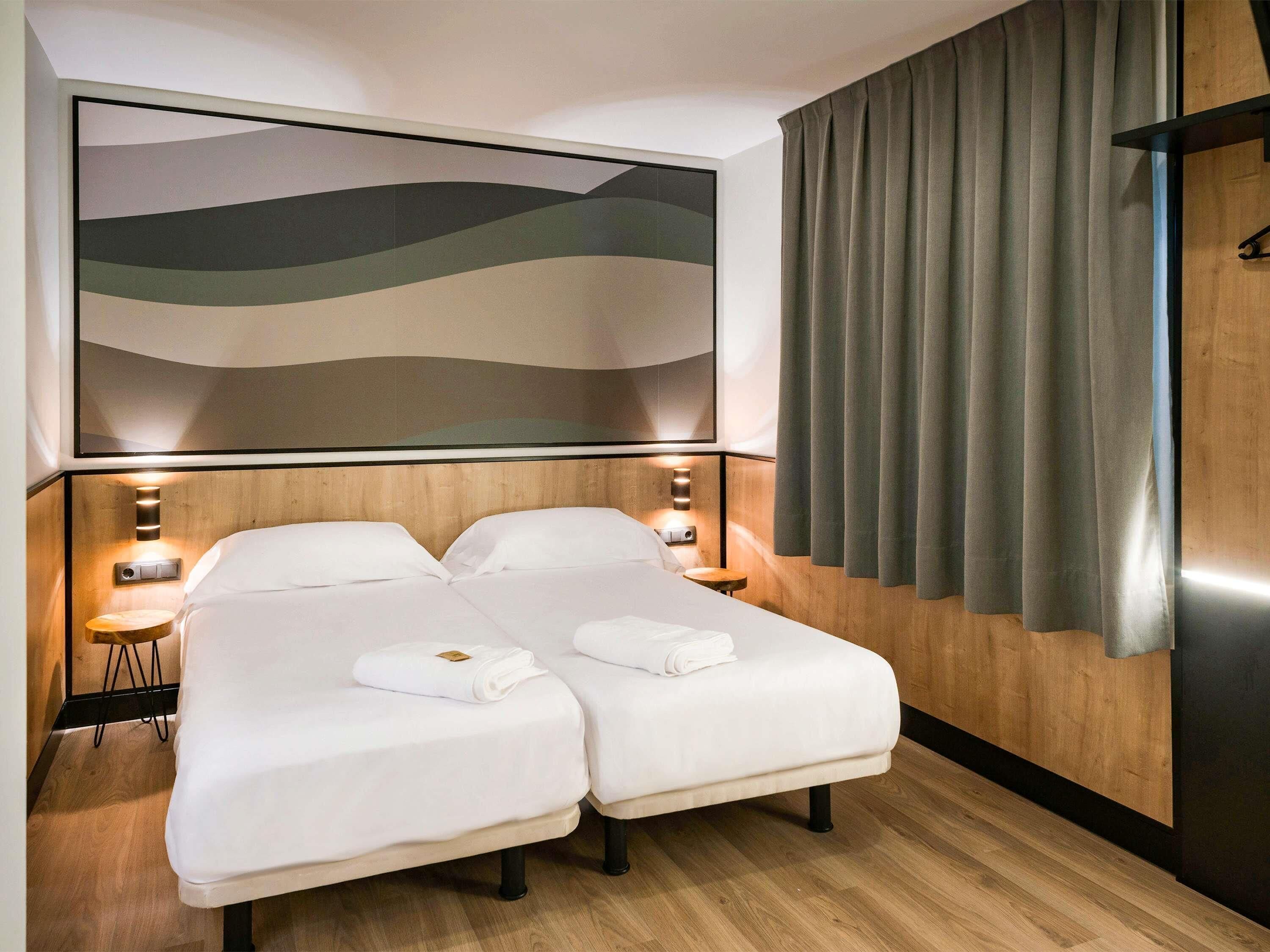 A hotel room with two beds, wooden headboard, a large abstract wall art, a wooden floor, and a curtain on the window.
