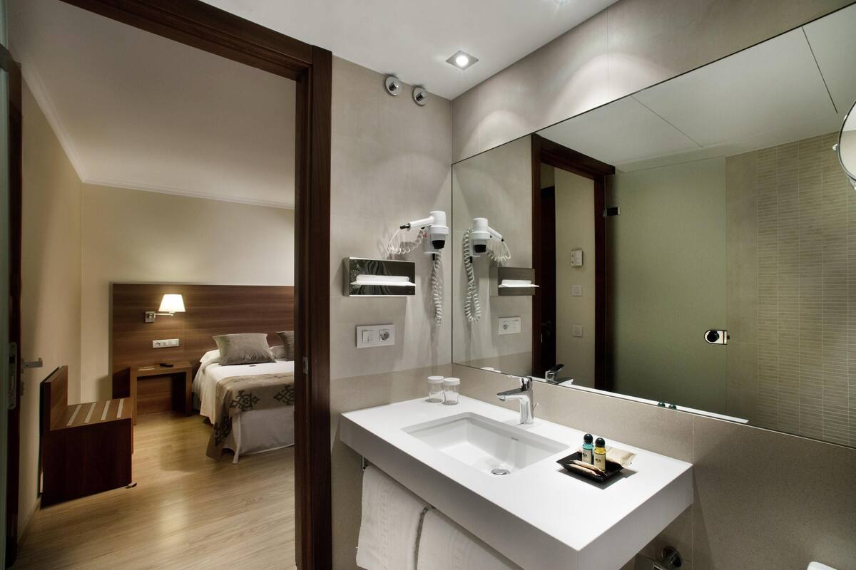 A modern hotel bathroom with a large mirror, a white sink, and a hairdryer on the wall.