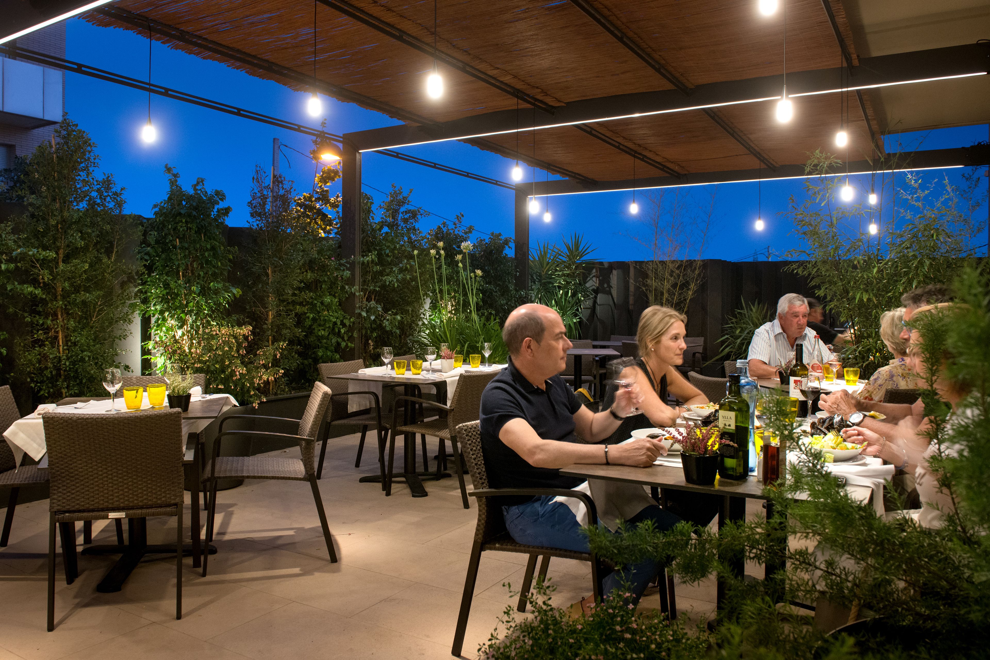 An outdoor dining area with tables and chairs, surrounded by greenery and under a covered roof.