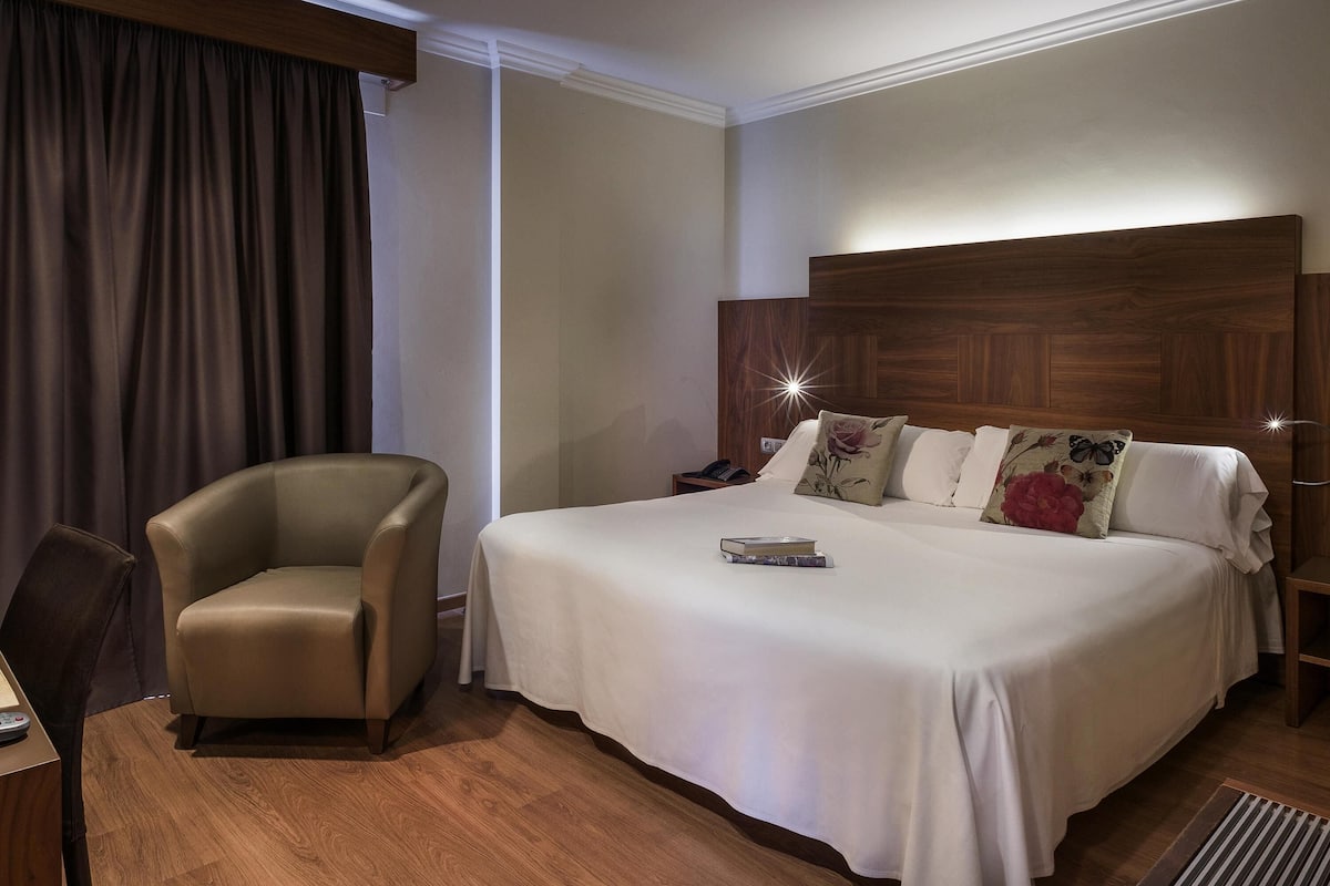 A hotel room with a large bed, a chair, a bedside table, and a wooden headboard.