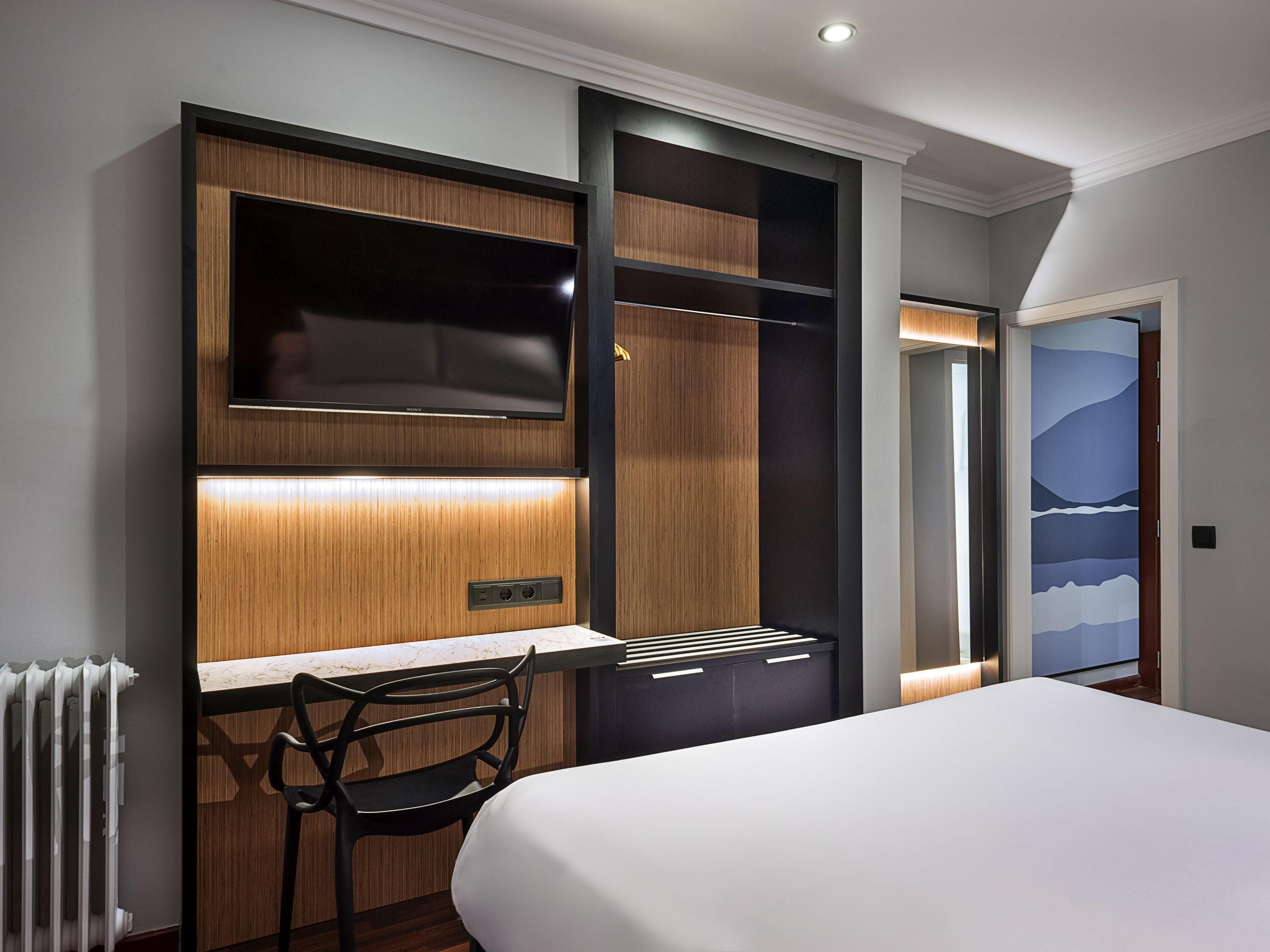 A modern hotel room with a bed, a desk with a chair, a TV, and a wardrobe.