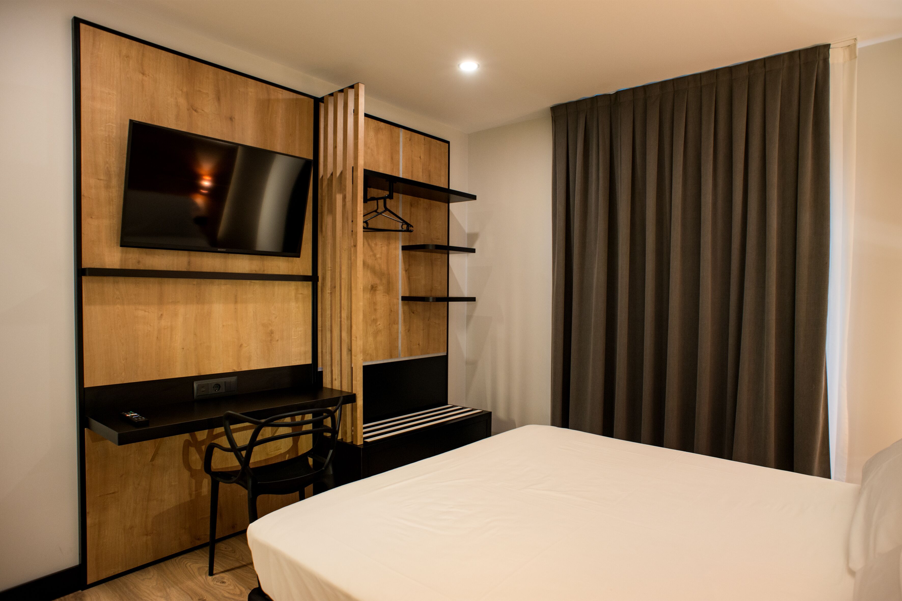 A modern hotel room with a bed, a desk, a TV mounted on a wooden panel, and a wardrobe with shelves.