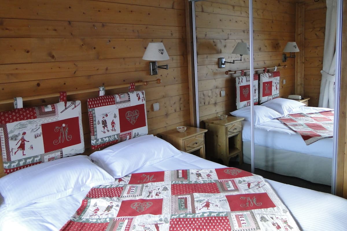 Une pièce chaleureuse aux murs en bois, avec un lit recouvert d’une couette rouge et blanche, deux tables de chevet dotées de lampes et un miroir qui reflète la pièce.