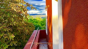 Un estrecho pasillo exterior con suelo de baldosas rojas, barandilla metálica y vistas a un paisaje verde bajo un cielo azul.