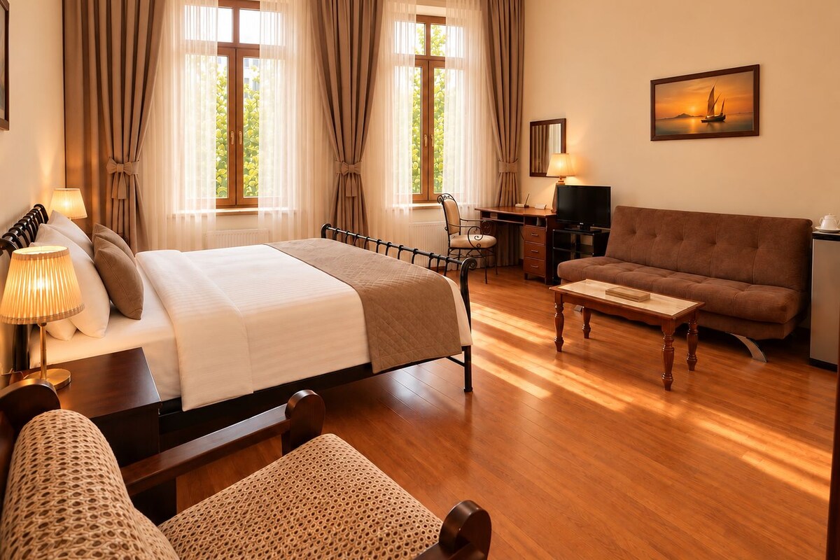A hotel room with a bed, a sofa, a desk with a TV, and a painting on the wall.