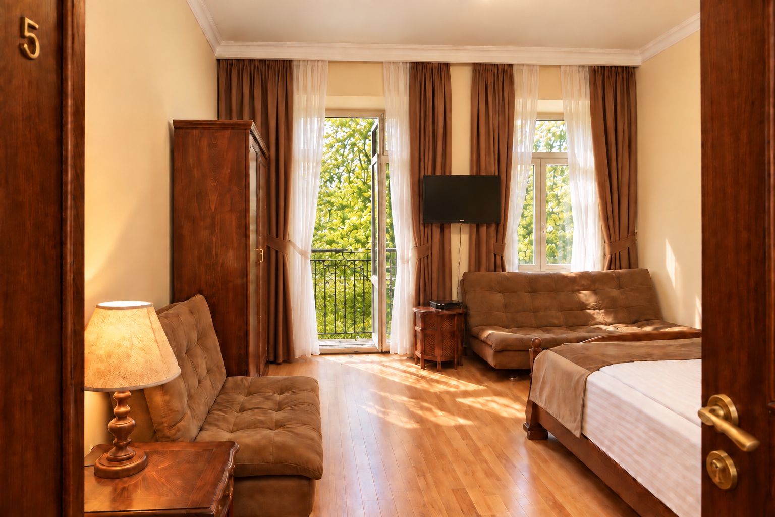 A hotel room with a bed, a sofa, a TV, a lamp, and a view of greenery outside.