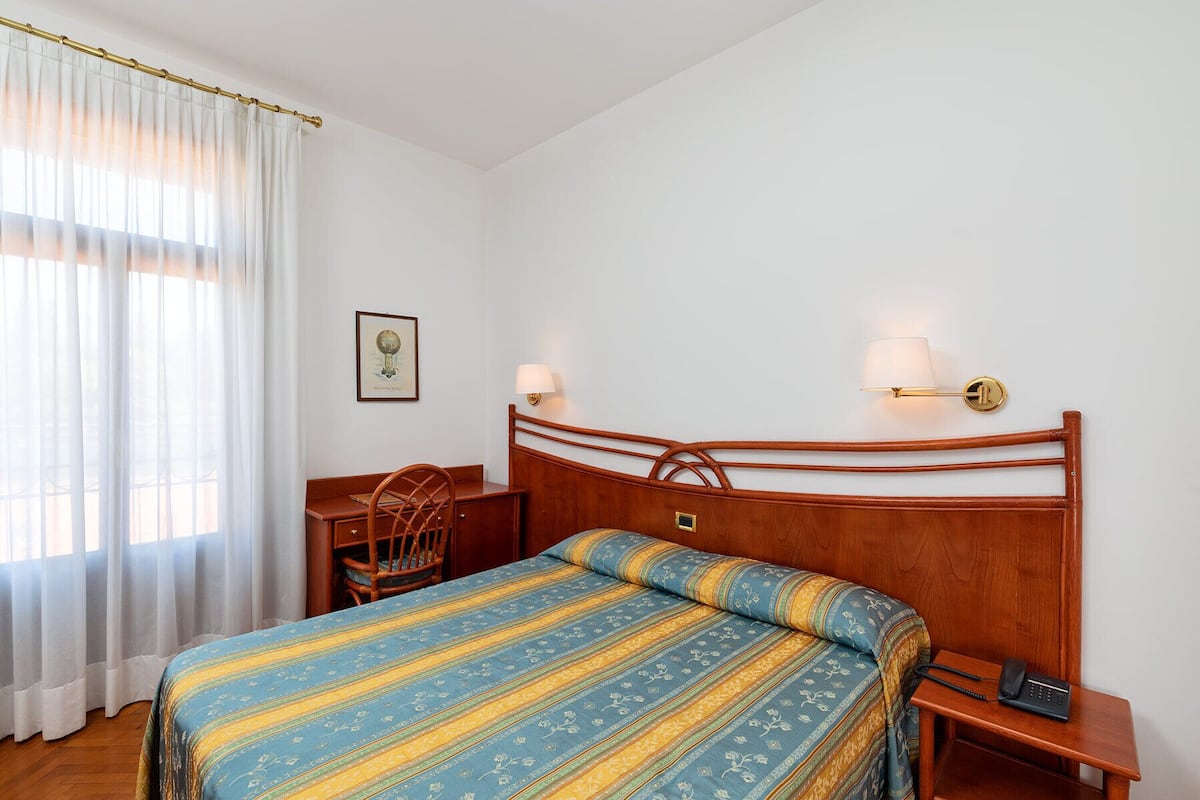Ein Hotelzimmer mit einem Holz-Kopfteil, einem Bett mit gestreifter Bettdecke, einem Schreibtisch mit Stuhl, zwei Wandlampen und einem gerahmten Bild an der Wand.