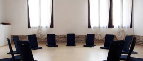 A room with eight black exercise chairs arranged in a circle, a pair of shoes on the floor, and a brick wall with a white curtain.