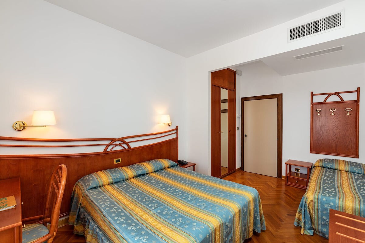 Ein Hotelzimmer mit zwei Betten, einem Holzkopfende, einem Nachttisch, einem Sessel, einem Kleiderschrank und einem Spiegel.