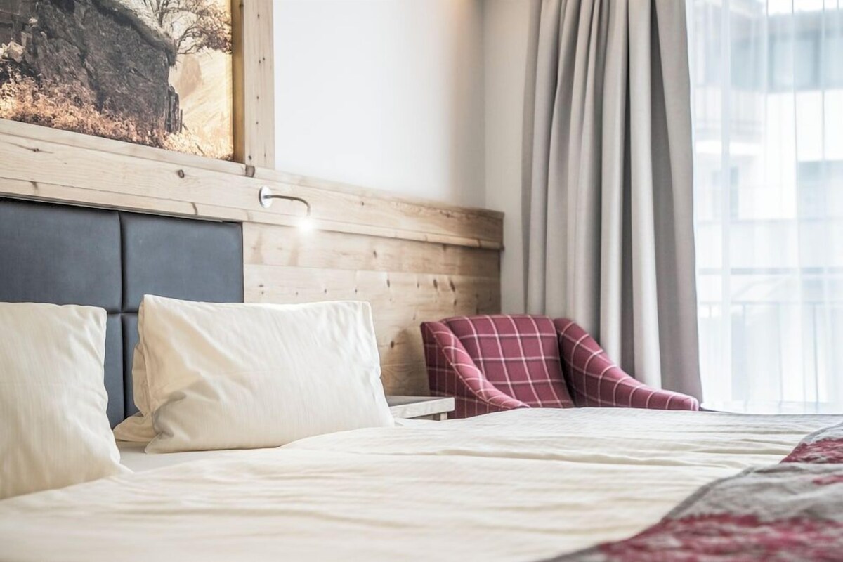 Habitación de hotel con cama, almohadas, cabecero, una silla y ventana con cortinas.