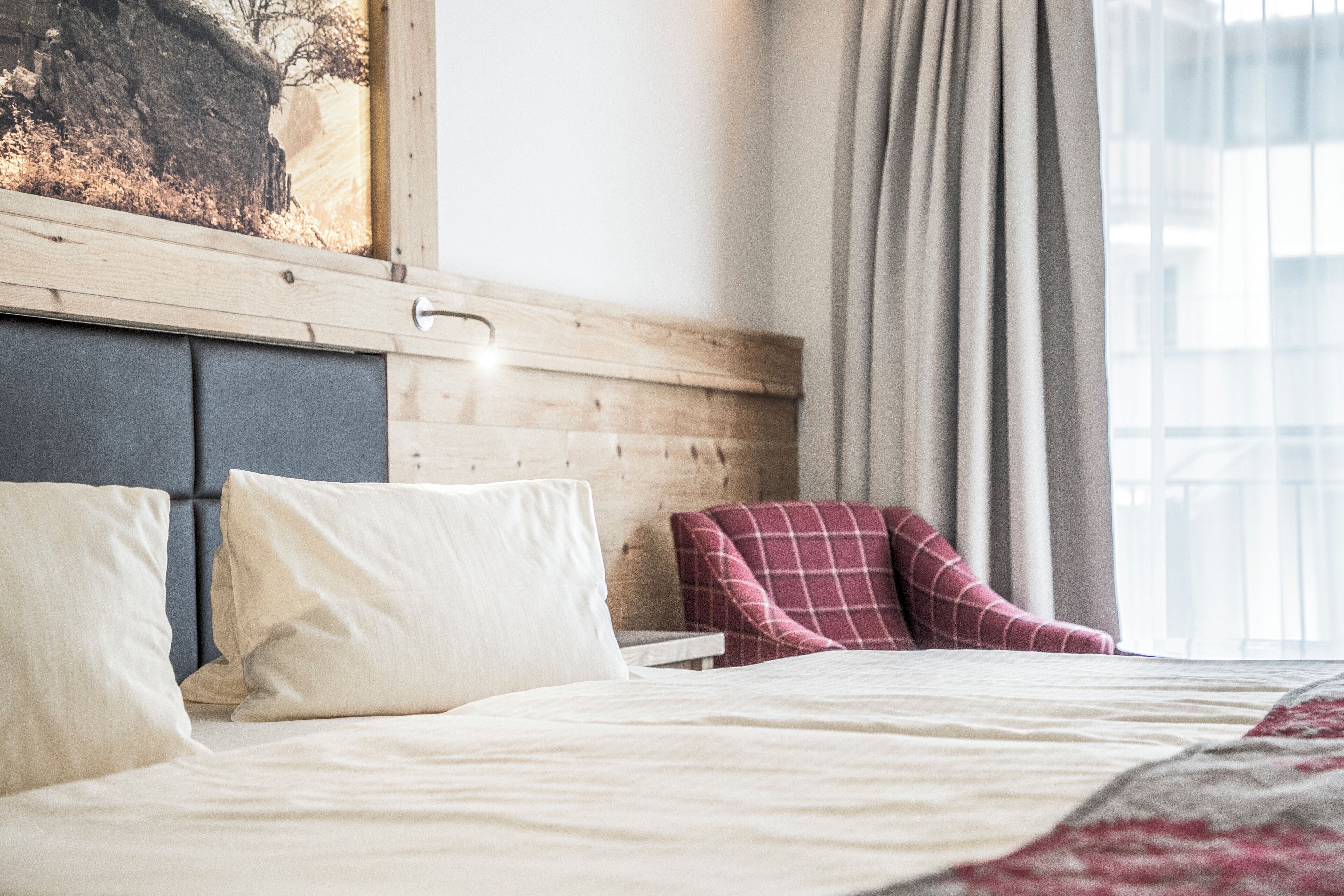Ein ordentlich bezogenes Bett mit Kissen, ein hölzernes Kopfteil, ein roter Karo-Sessel und ein Fenster mit Vorhängen.