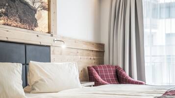 Ein ordentlich bezogenes Bett mit Kissen, ein hölzernes Kopfteil, ein roter Karo-Sessel und ein Fenster mit Vorhängen.