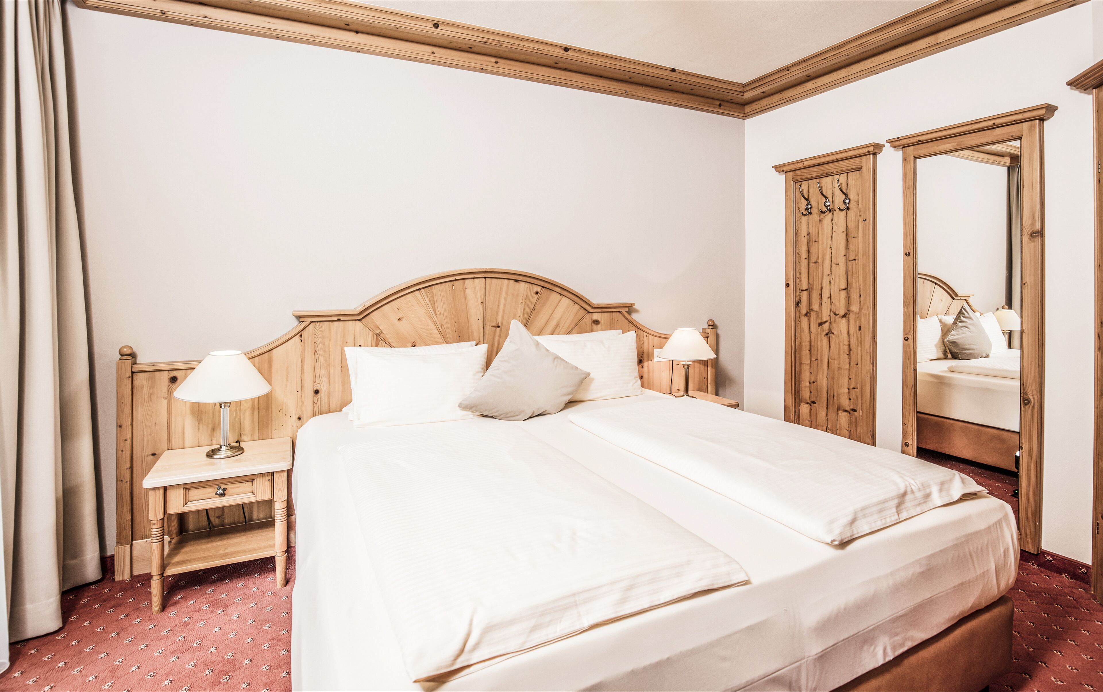 Habitación de hotel con cama de madera, dos mesitas de noche con lámparas, un espejo grande y una alfombra roja estampada.