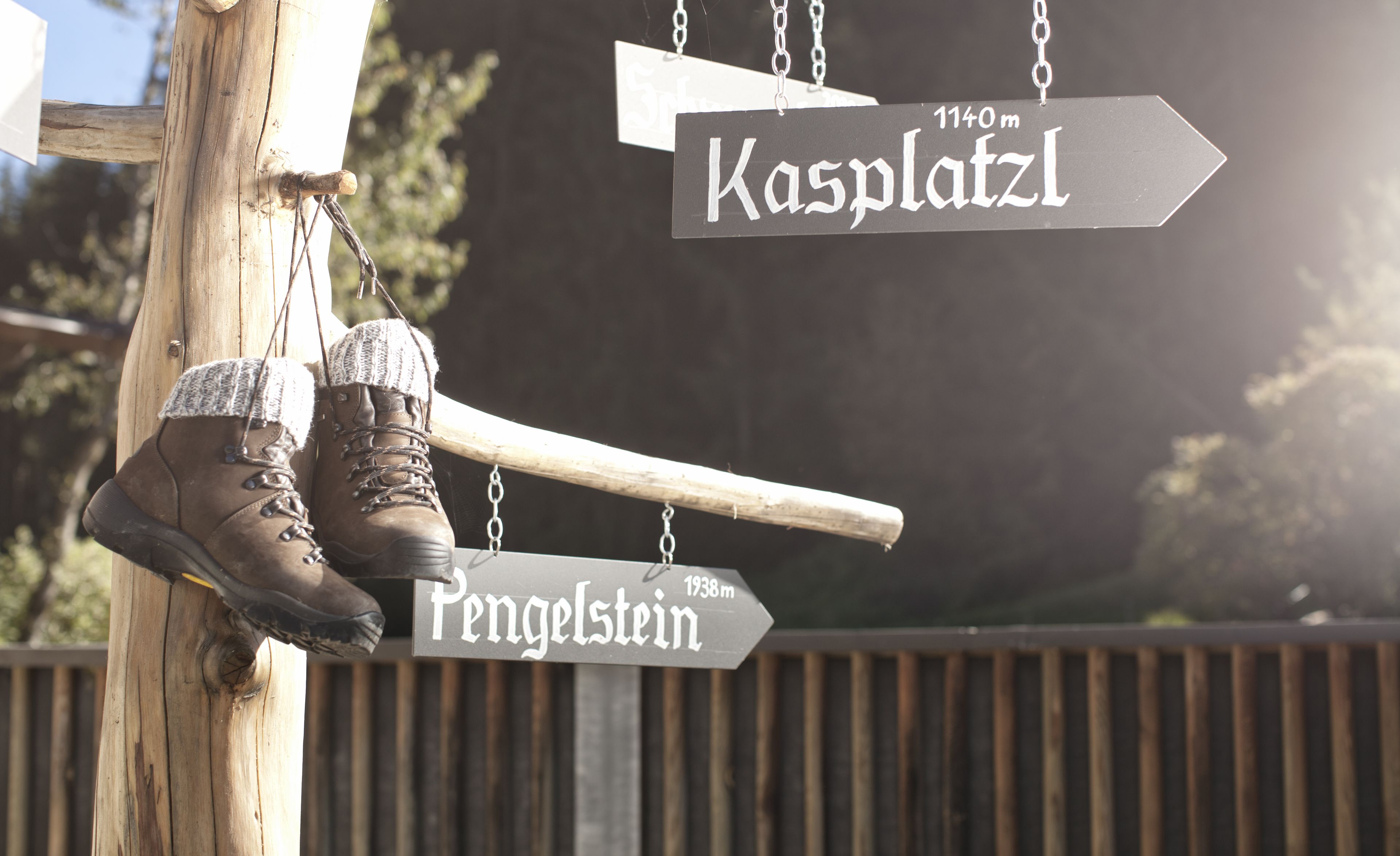 Un cartel de madera con indicaciones direccionales hacia 'Kasplatz' y 'Pengelstein', mostrando distancias de 1140m y 1938m respectivamente. Un par de botas cuelga del cartel.