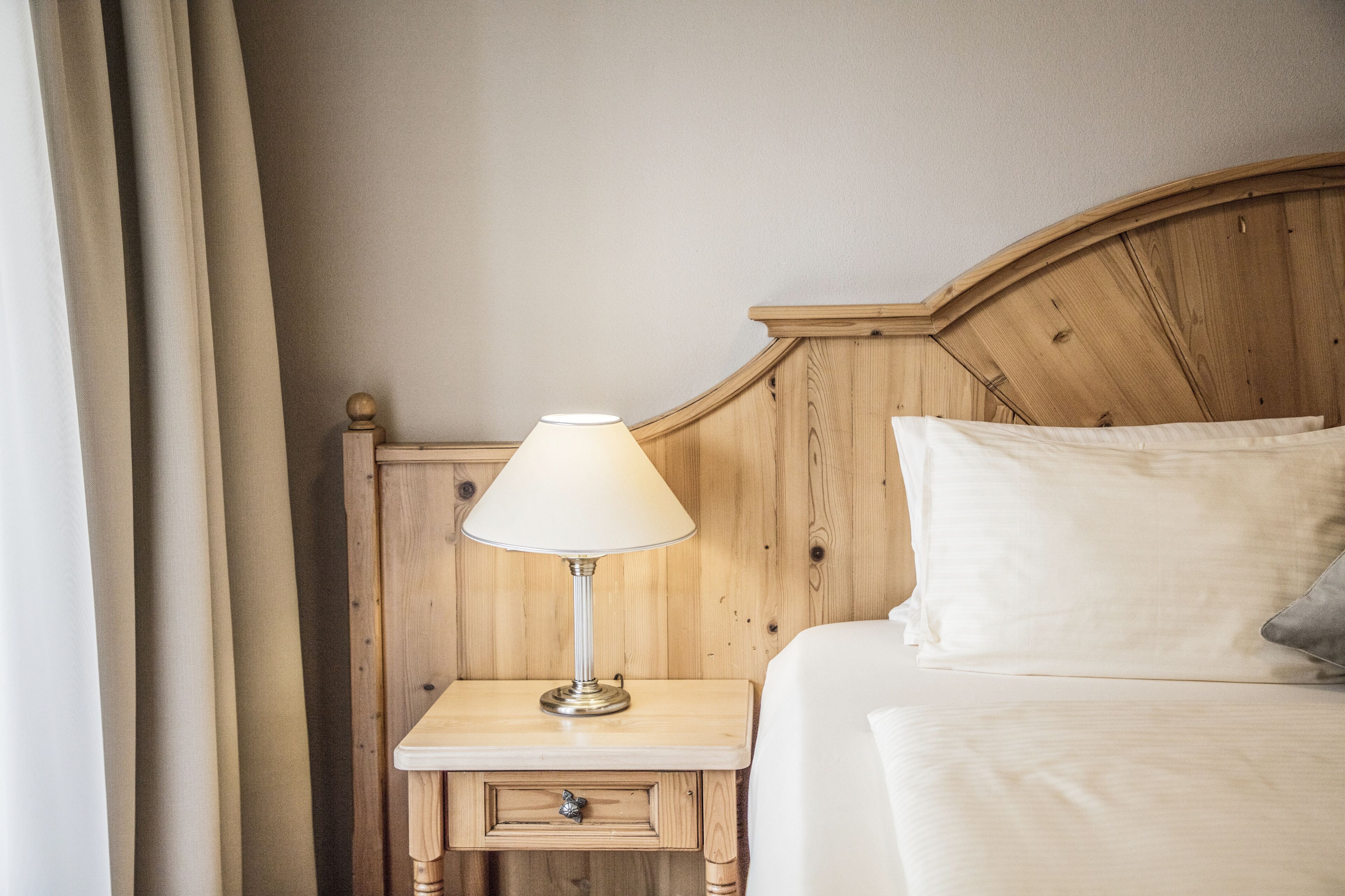 Una cama bien tendida con cabecera de madera, una mesita de noche con lámpara y un cajón pequeño.