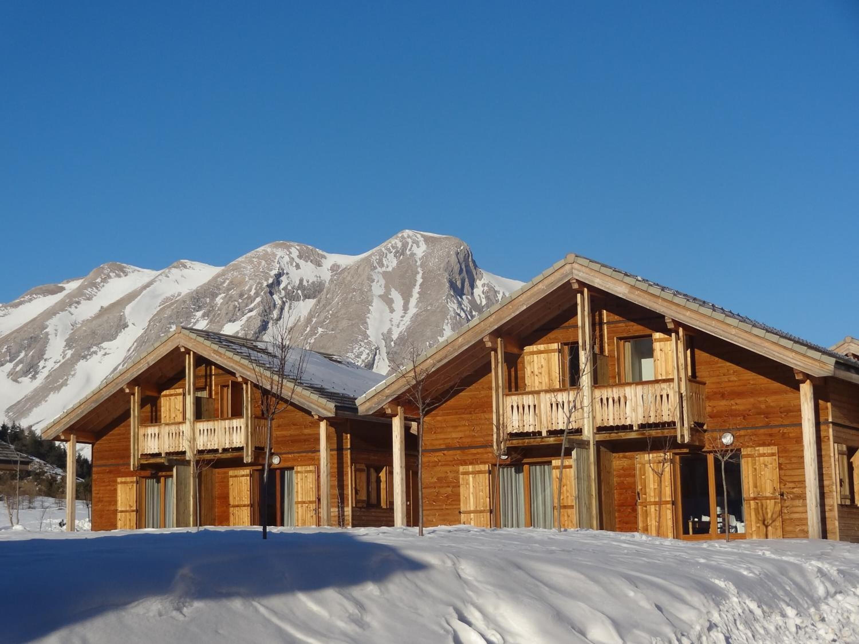 A wooden chalet with a balcony, set against a snowy mountain backdrop.