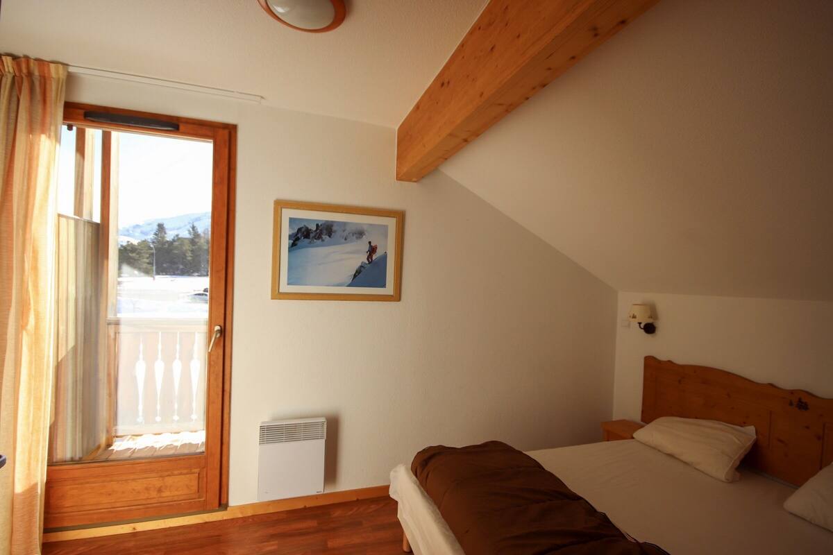Ein Schlafzimmer mit Holzbalkendecke, einem Bett mit braunem Kopfteil, einem Nachttisch, einer Wandlampe und einem gerahmten Bild einer verschneiten Landschaft.